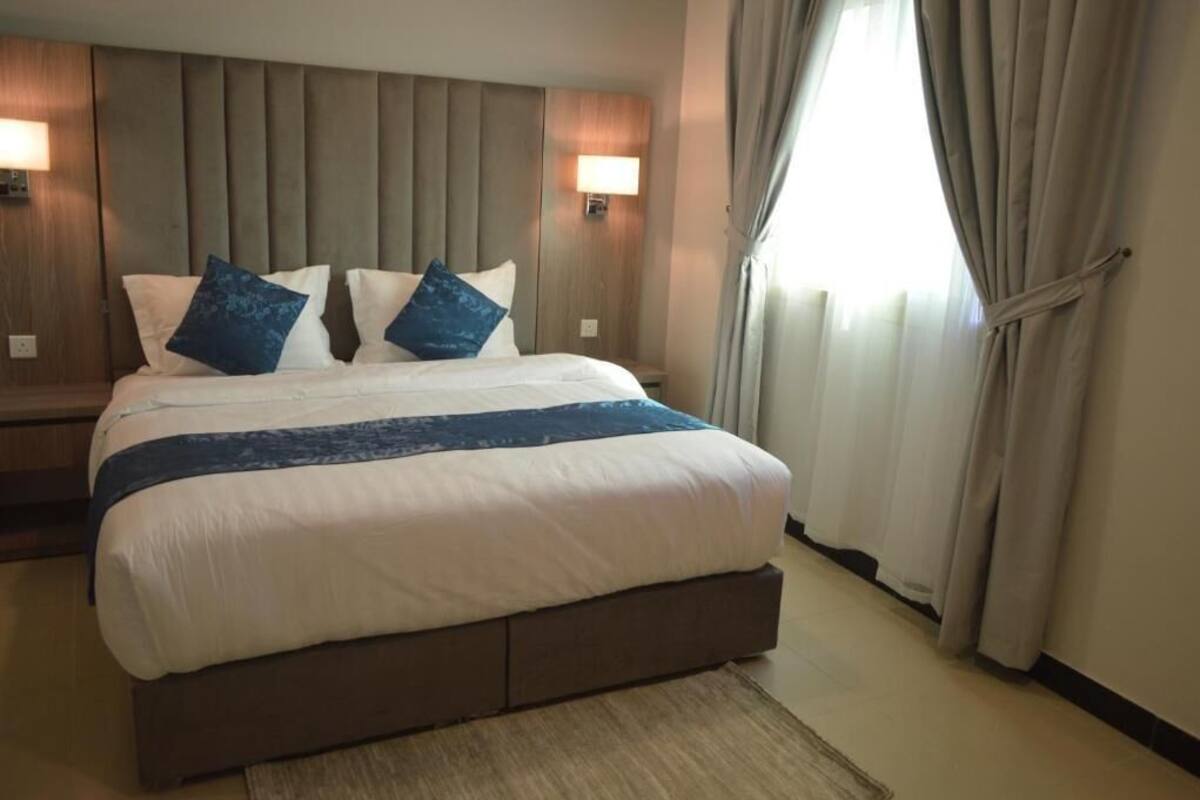 A hotel room with a large bed, two wall-mounted lamps, a window with curtains, and a wooden headboard.