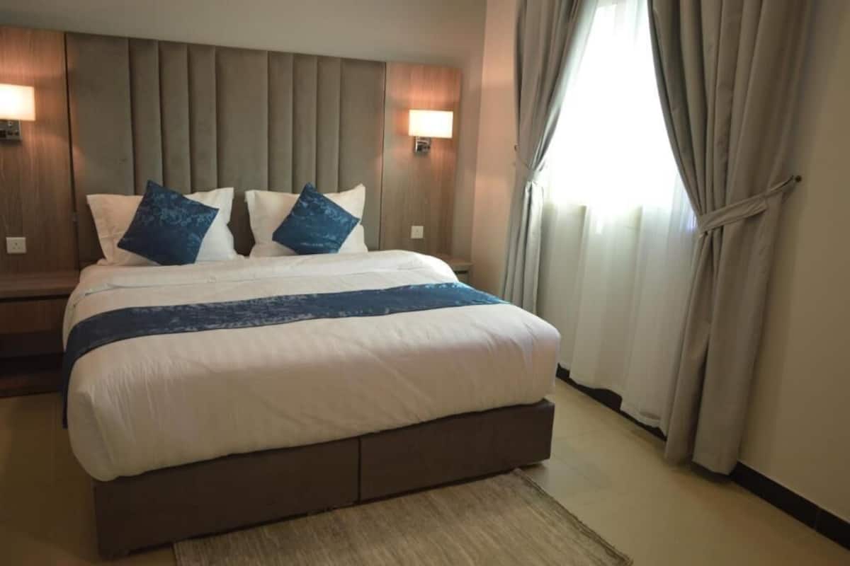 A hotel room with a large bed, two bedside lamps, a headboard, and a window with curtains.