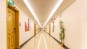 Un pasillo largo e iluminado, con una puerta de madera a la izquierda, una planta en maceta a la derecha y cuadros en las paredes.