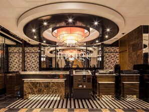 Un hall d’hôtel moderne, agrémenté d’un lustre central, d’un comptoir de réception en marbre et de motifs décoratifs sur les murs.