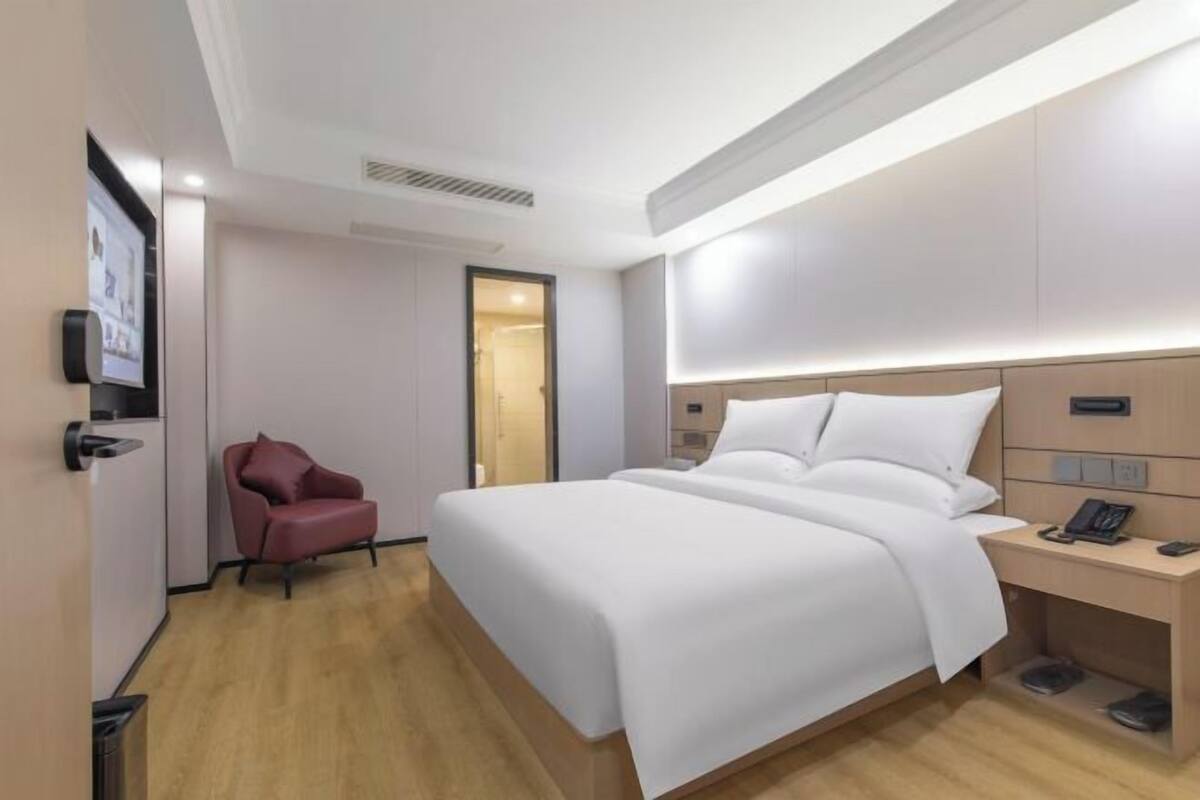 Een hotelkamer met een groot bed, een nachtkastje met een telefoon, een rode stoel en een deur naar een badkamer.