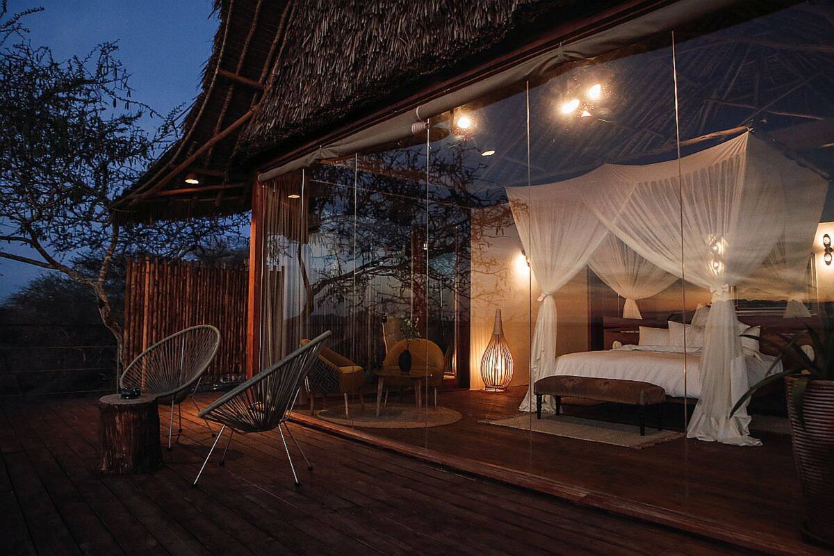 Une chambre à coucher aux murs de verre, dotée d’un lit à baldaquin, d’une terrasse en bois et d’un espace salon extérieur.