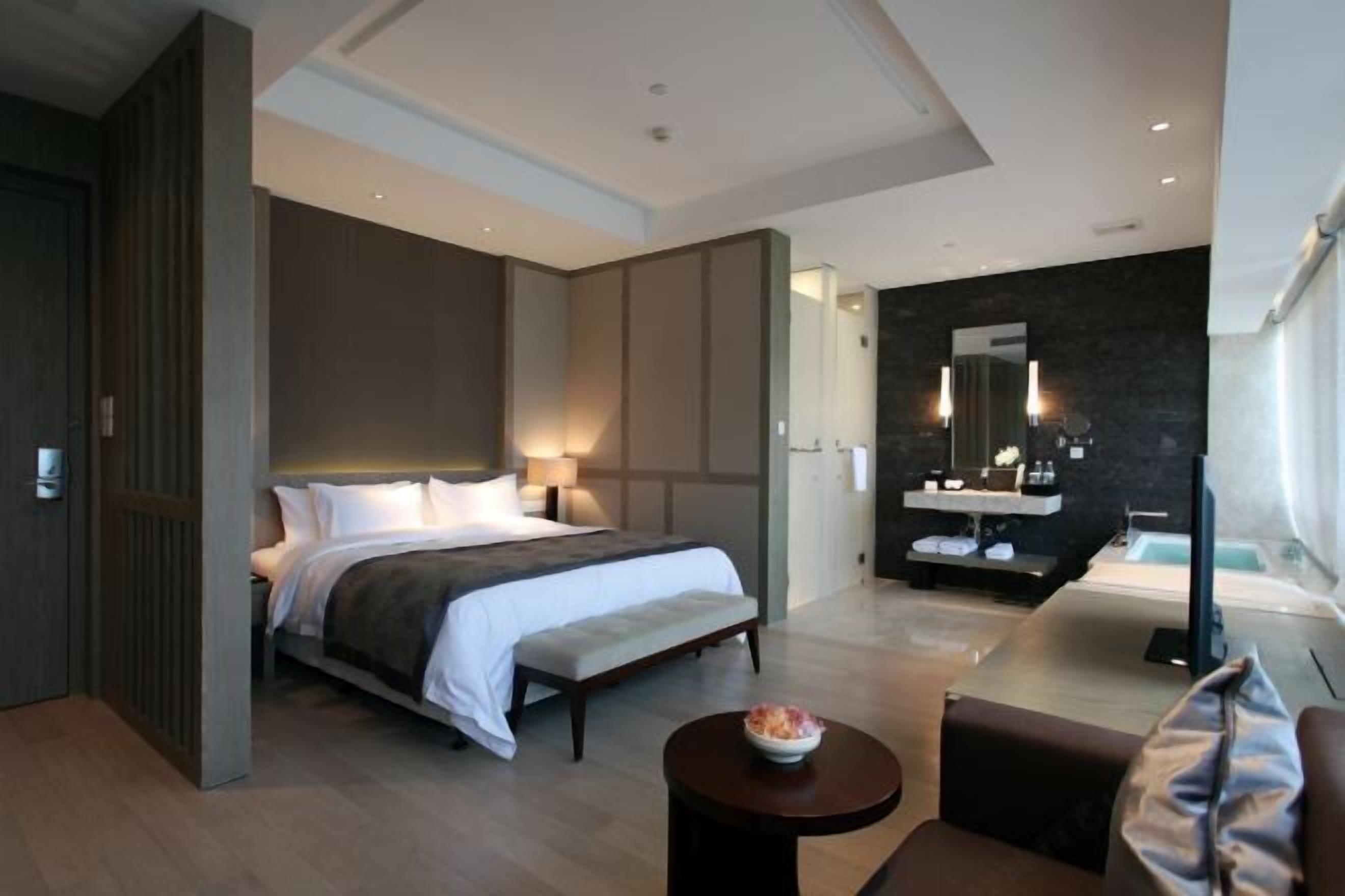 A modern hotel room with a large bed, bedside tables, a small round table with a bowl, a sofa, a bathroom with a mirror, and a TV.