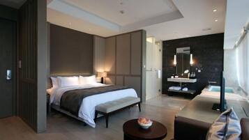 A modern hotel room with a large bed, bedside tables, a small round table with a bowl, a sofa, a bathroom with a mirror, and a TV.