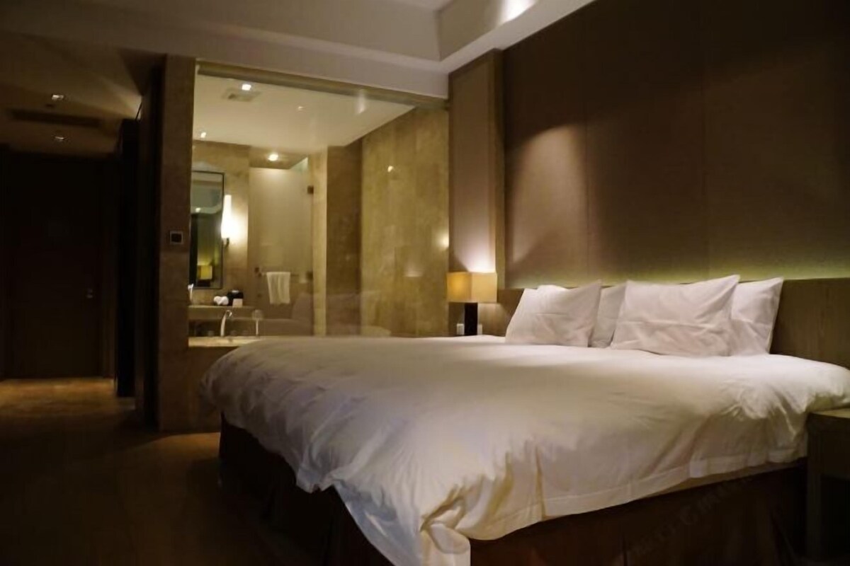 A modern hotel room with a large bed, a bedside table, a mirror, and a bathroom visible through an open door.