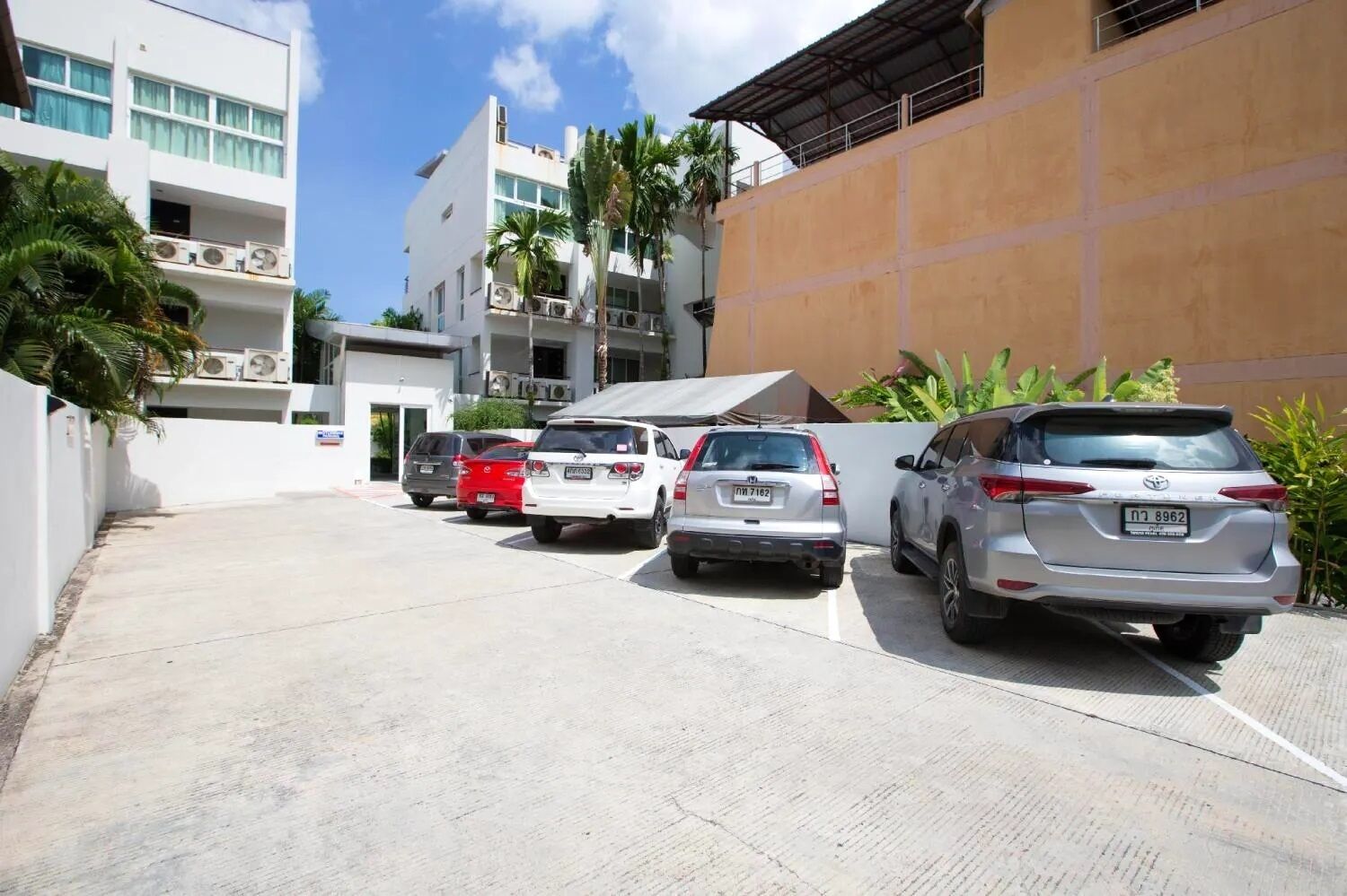 Un espace de stationnement avec plusieurs voitures, un immeuble à étages doté de balcons et quelques espaces verts.