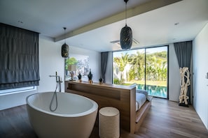 A modern bathroom with a freestanding bathtub, a wooden vanity, and a large window overlooking a pool and greenery. - Cocoon villas by Lofty (Sa Khu)