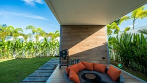 Ein moderner Außenbereich mit gebogenen Sitzgelegenheiten und orangefarbenen Kissen, umgeben von einer Holzterrasse und üppigem Grün.