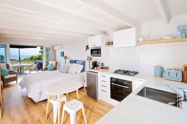 A modern kitchen with a central island, a dining area with a white table, and a bedroom with a large bed and a view of the outdoors. - Pointbreak Studio (Te Awanga)