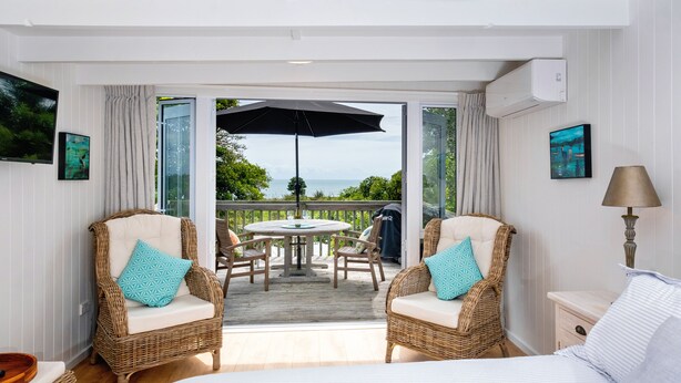 A bedroom with a bed, two wicker chairs with cushions, a small bedside table, and a lamp. There is a balcony with a table, chairs, and an umbrella. - Pointbreak Studio (Te Awanga)