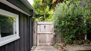 A wooden pathway leading to a gated entrance surrounded by lush greenery and a wooden deck. - Pointbreak Studio (Te Awanga)