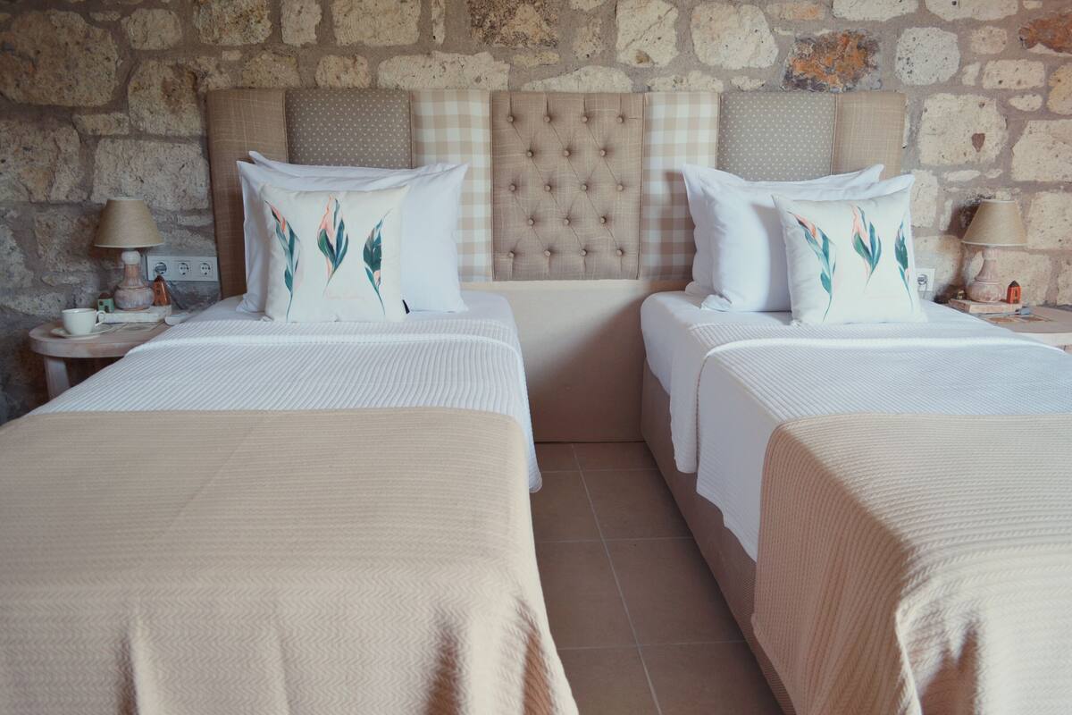 A hotel room with two beds, each with a headboard, a nightstand with a lamp, and a wall with a stone pattern.