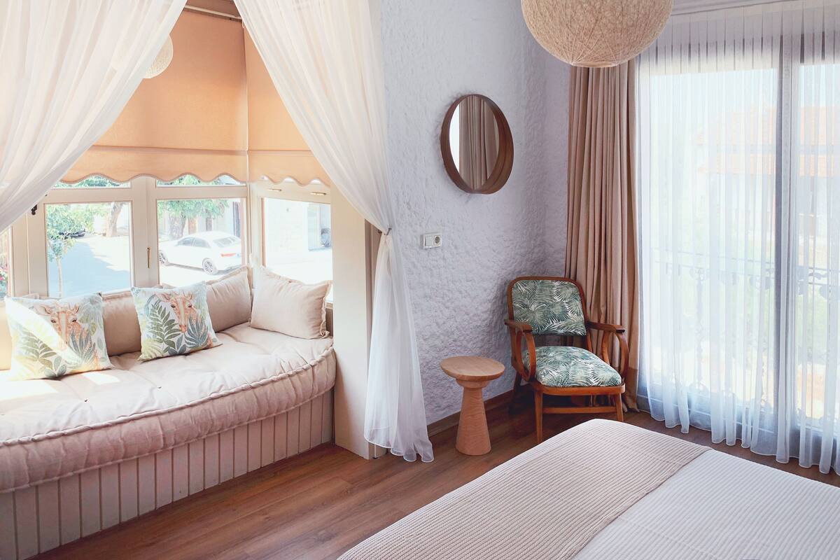A bedroom with a bed, a chair, a small table, a mirror, and a window with curtains.