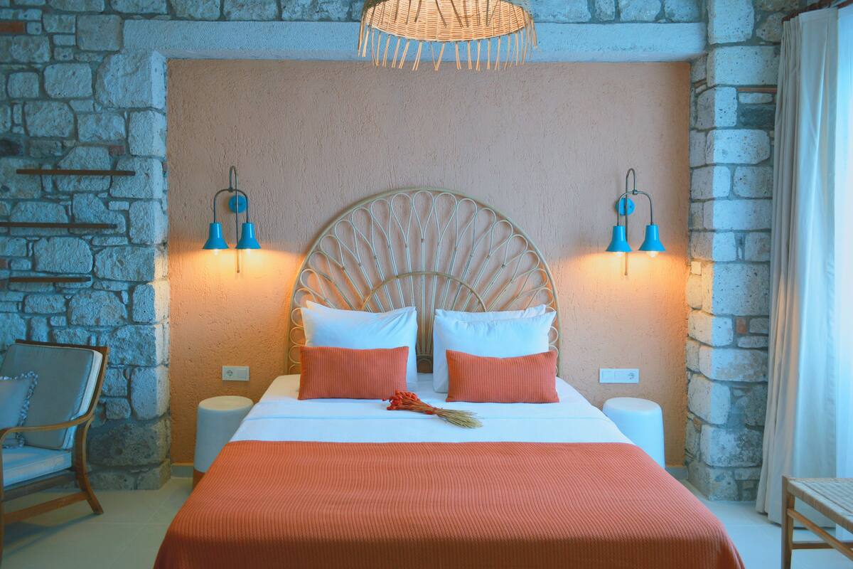 A bedroom with a stone wall, a large bed with white and orange bedding, two bedside lamps, and a wicker headboard.