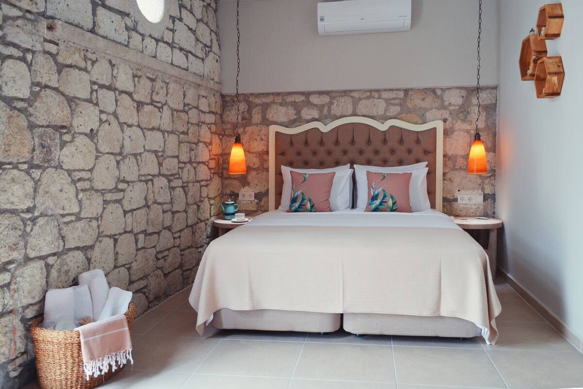 A bedroom with a stone wall, a tufted headboard, two bedside tables with lamps, a basket of towels, and a bed with a white comforter.
