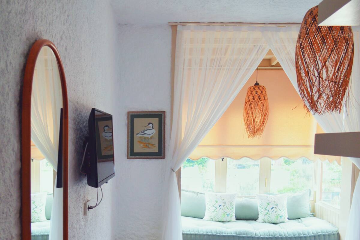 A bedroom with a bed, a yellow ottoman, a large mirror, and a window with curtains.