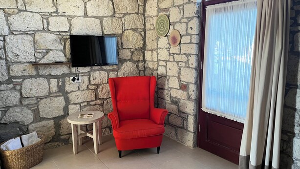 A room with a stone wall, a red armchair, a small round table, a flat-screen TV mounted on the wall, and a window with sheer curtains. - Rijeka Alaçatı (Cesme)