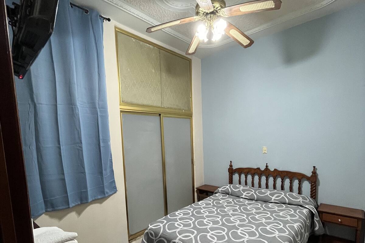 Ein Schlafzimmer mit einem Bett, einem Deckenventilator, einem Holzstuhl, einem kleinen Tisch und einem an der Wand montierten Fernseher.