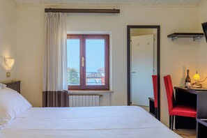 Una habitación de hotel con cama, escritorio con silla, ventana con cortinas, radiador y lámpara de pared.