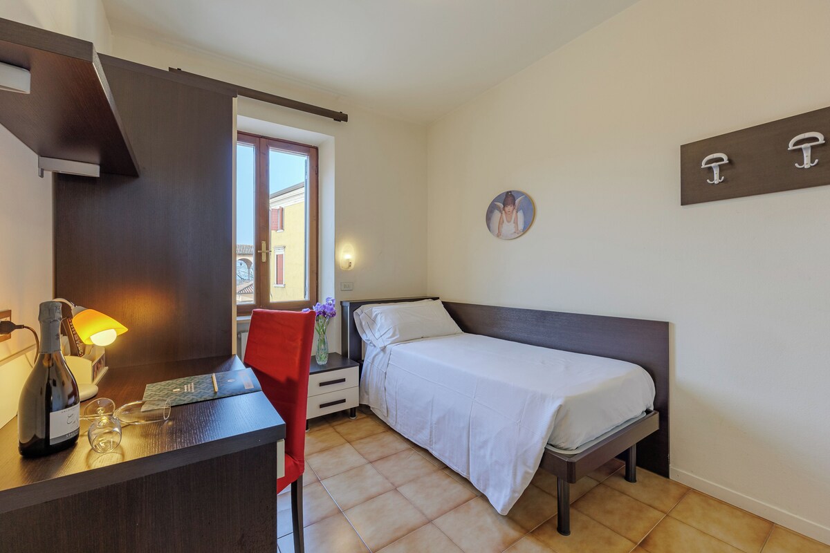 Un dormitorio con cama, escritorio con lámpara, silla y estante con ganchos.
