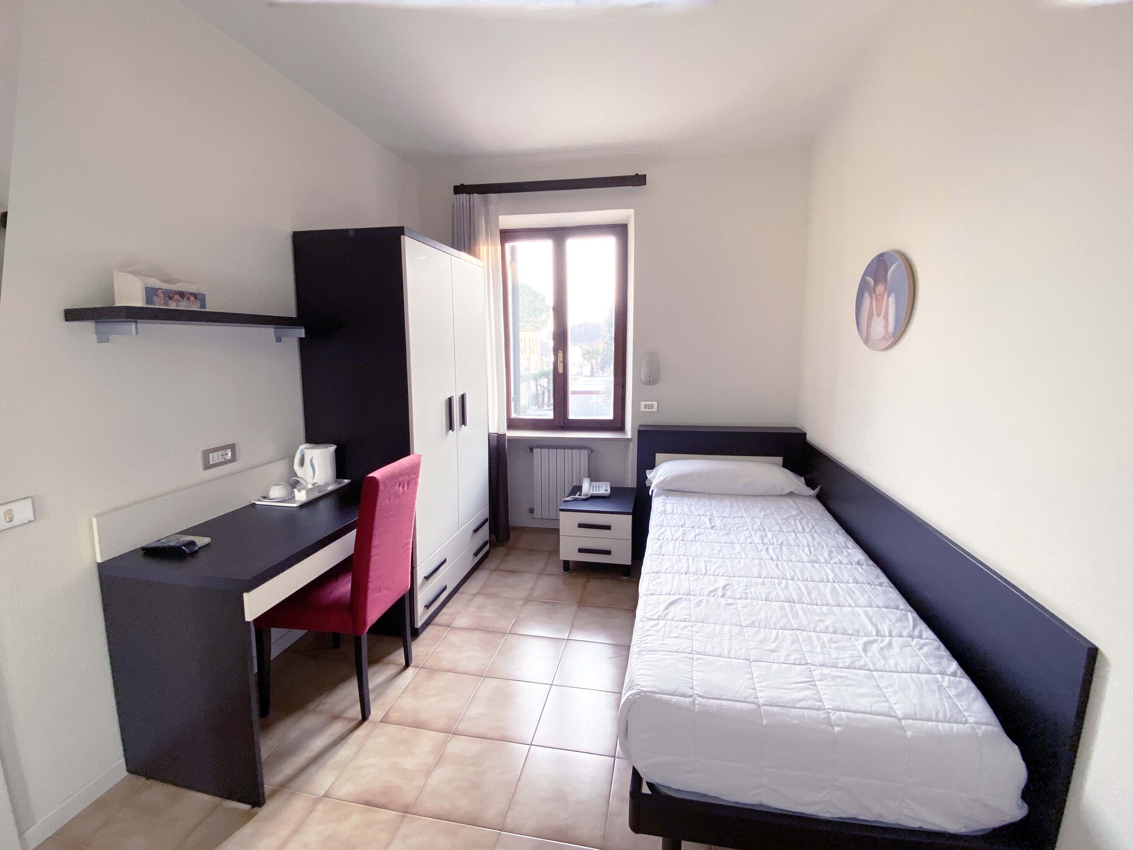 A small, well-lit room with a bed, a desk, and a chair. There is a window providing natural light, and a radiator under the window.