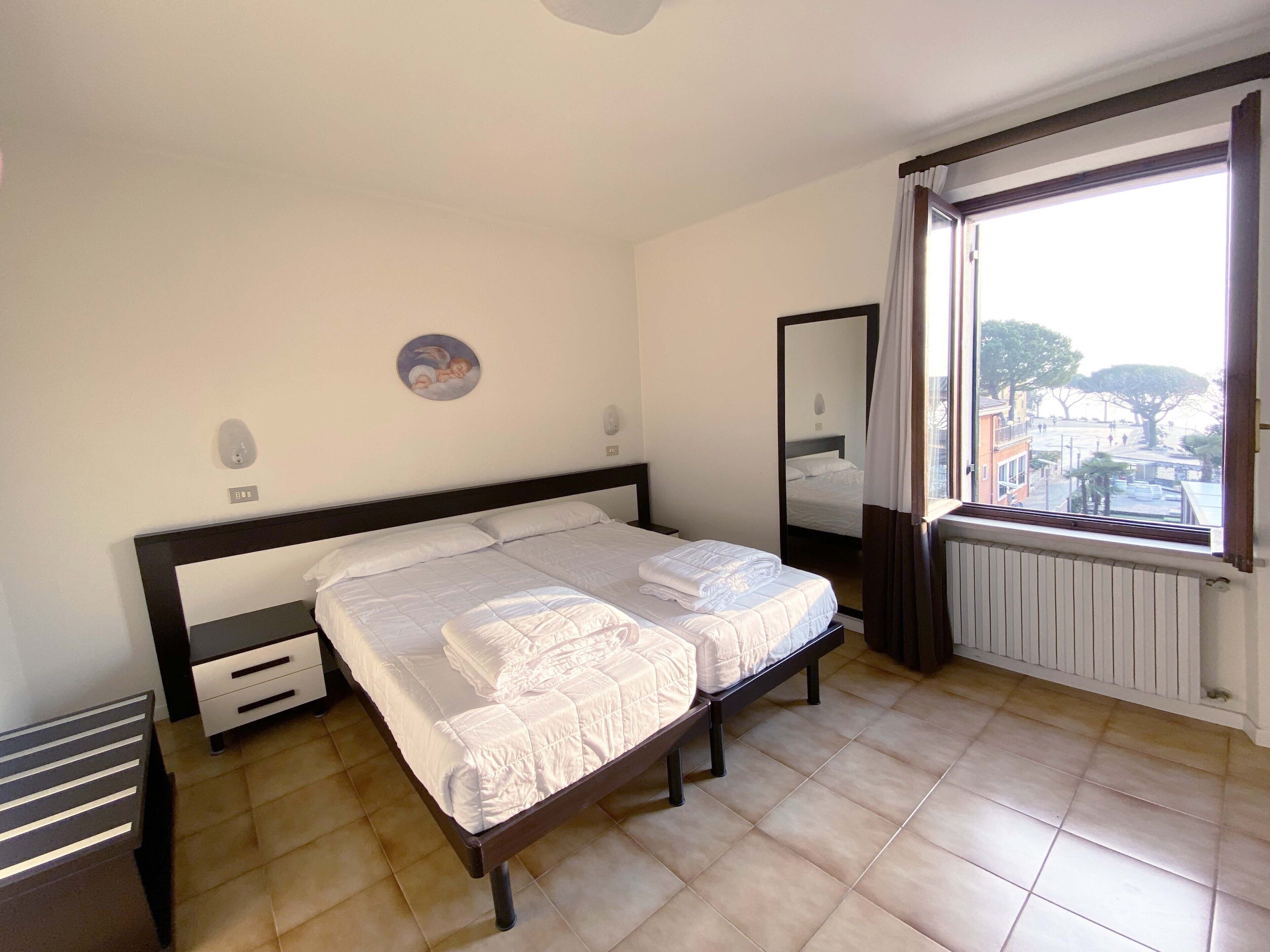 A bedroom with a large bed, a bedside table, a mirror, and a window with a view of the city.