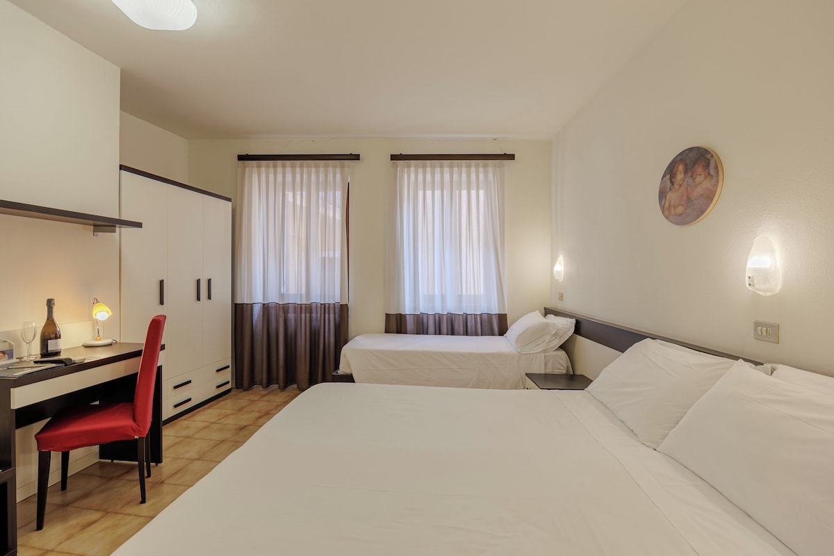Habitación de hotel con cama, escritorio con silla, dos ventanas con cortinas, un cuadro en la pared y mesita de noche con lámpara.