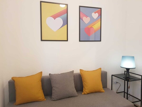 A room with a grey sofa, two mustard-colored pillows, and a small table with a lamp. Two framed abstract art pieces with heart shapes are on the wall.