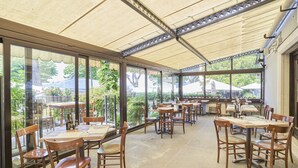 A covered outdoor dining area with wooden tables and chairs, large windows, and a view of greenery. - Hotel Astoria (Garda)