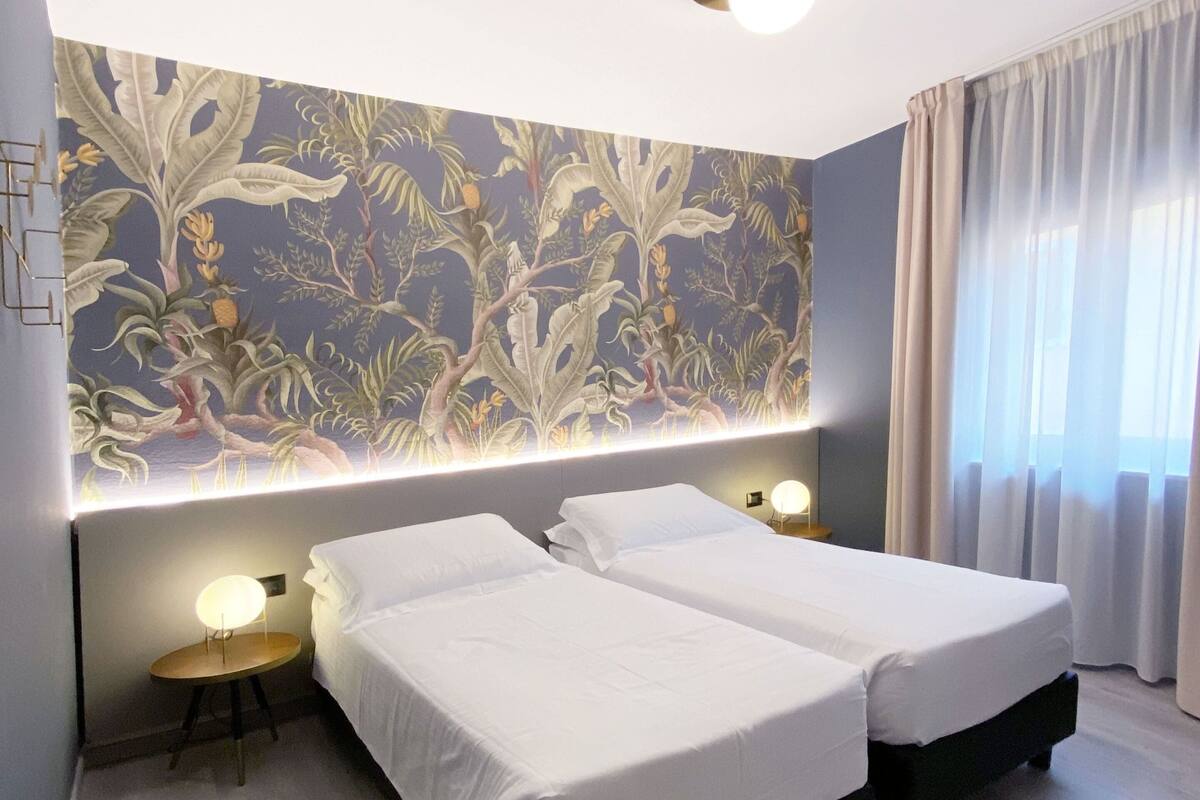 A hotel room with two beds, a floral wallpaper, a ceiling light, and a window with curtains.