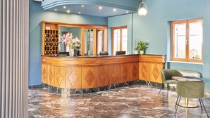A reception area with a marble floor, a wooden reception desk, a green chair, and a potted plant. - Hotel Astoria (Garda)