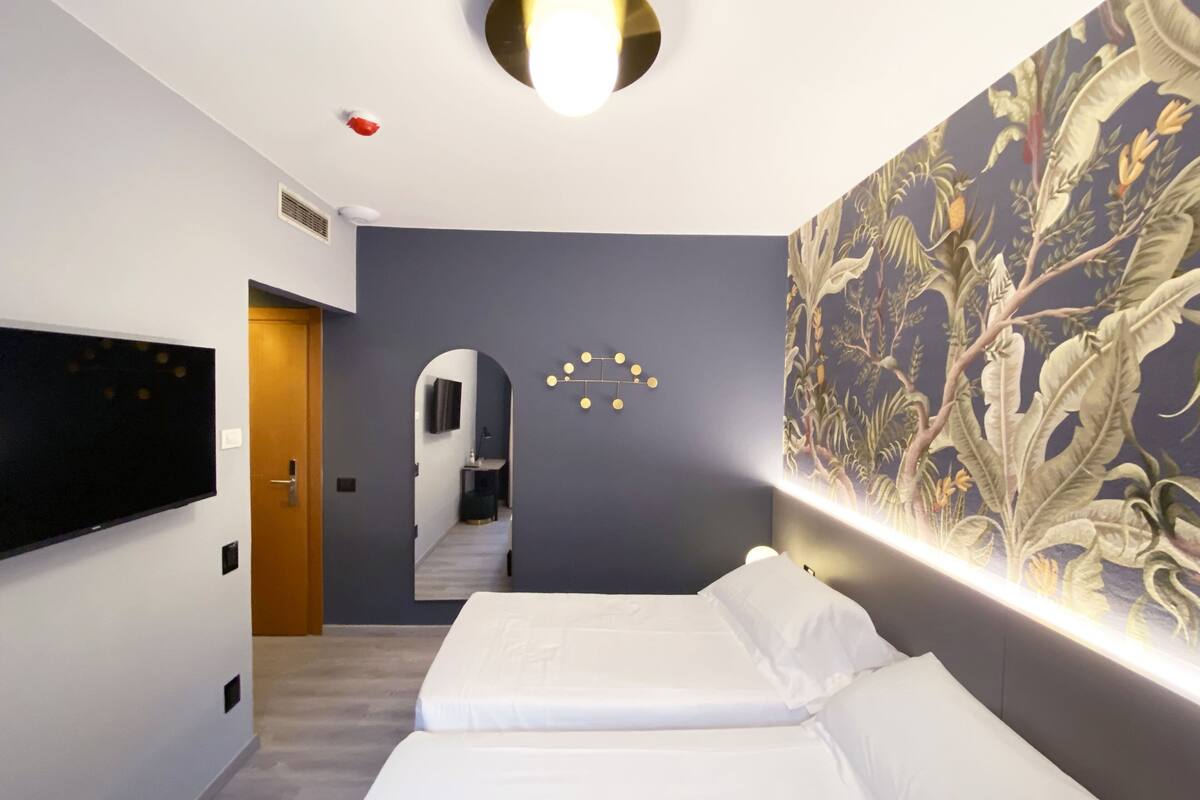 A modern hotel room with a large bed, a flat-screen TV mounted on the wall, a ceiling light fixture, and a decorative wall panel.
