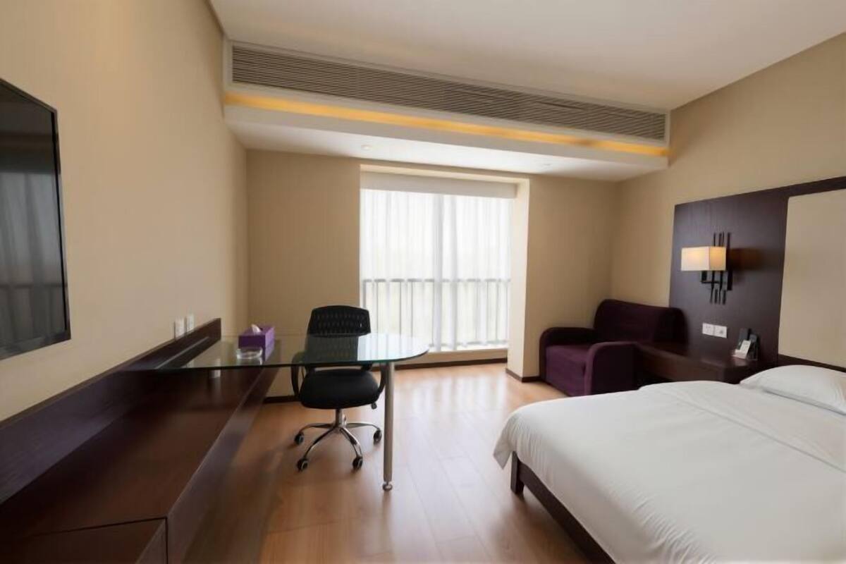 A hotel room with a bed, a desk with a chair, a sofa, a TV, and a window with blinds.