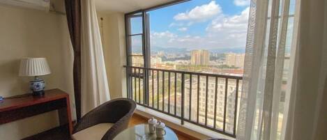 A balcony with wicker chairs and a small table, overlooking a cityscape with buildings and a clear sky.