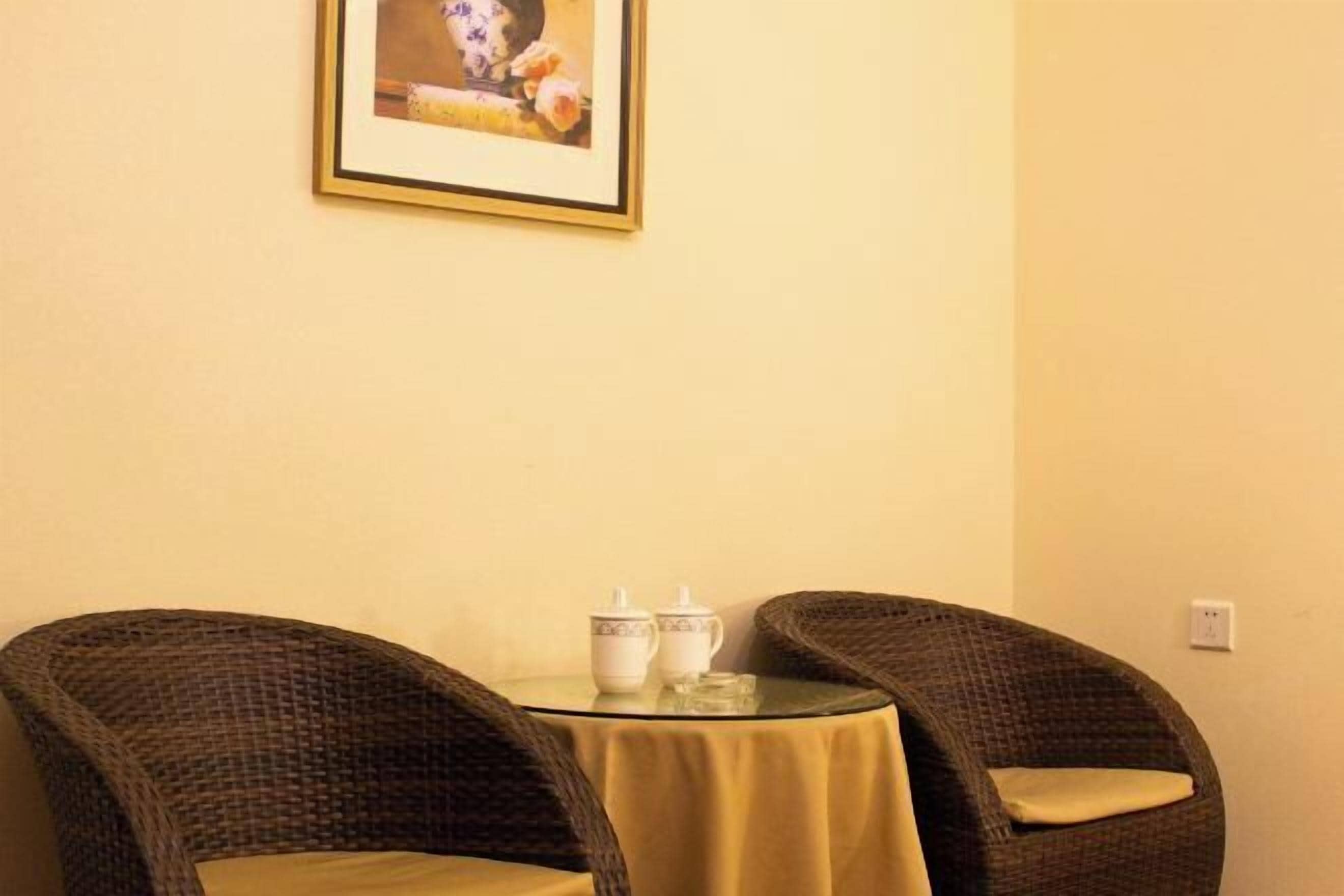 Una habitación con dos sillones de mimbre alrededor de una pequeña mesa redonda con mantel amarillo, dos tazas de cerámica blanca y un cuadro en la pared.