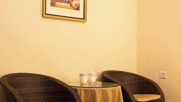 Una habitación con dos sillones de mimbre alrededor de una pequeña mesa redonda con mantel amarillo, dos tazas de cerámica blanca y un cuadro en la pared.