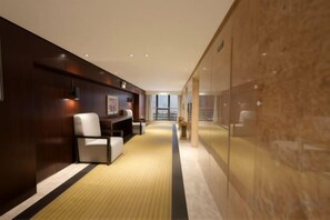 A hotel corridor with a carpeted floor, a seating area with chairs, and a glass wall reflecting the interior.