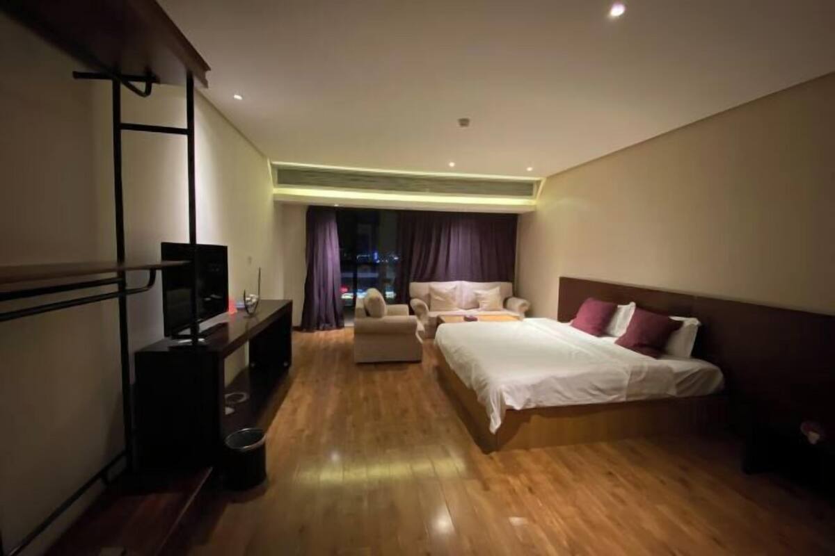 A modern hotel room with a wooden floor, a bed with white bedding, a sofa, a TV on a stand, and a window with curtains.