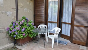 A patio with a glass table and two white chairs, a potted plant with purple flowers, and a door with wooden shutters. - Poolside Stay in Lazise (Lazise)