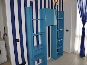 A room with a blue shelving unit against a striped wall, a washing machine, and a window with curtains.
