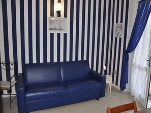 Ein Zimmer mit einem blauen Sofa, einem kleinen Tisch, einer Wandregal mit einem Bild und einem Vorhang.