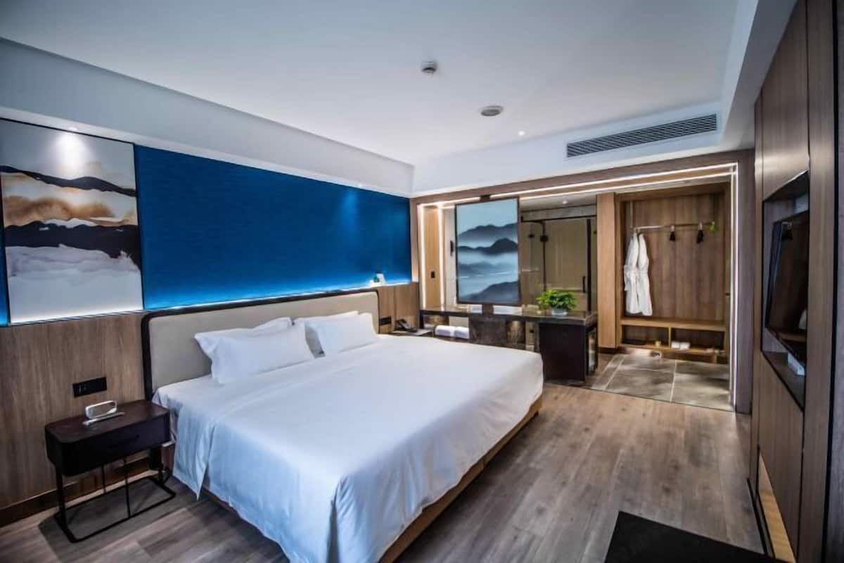 A modern hotel room with a large bed, a bedside table, a desk with a chair, a large mirror, and a wardrobe.