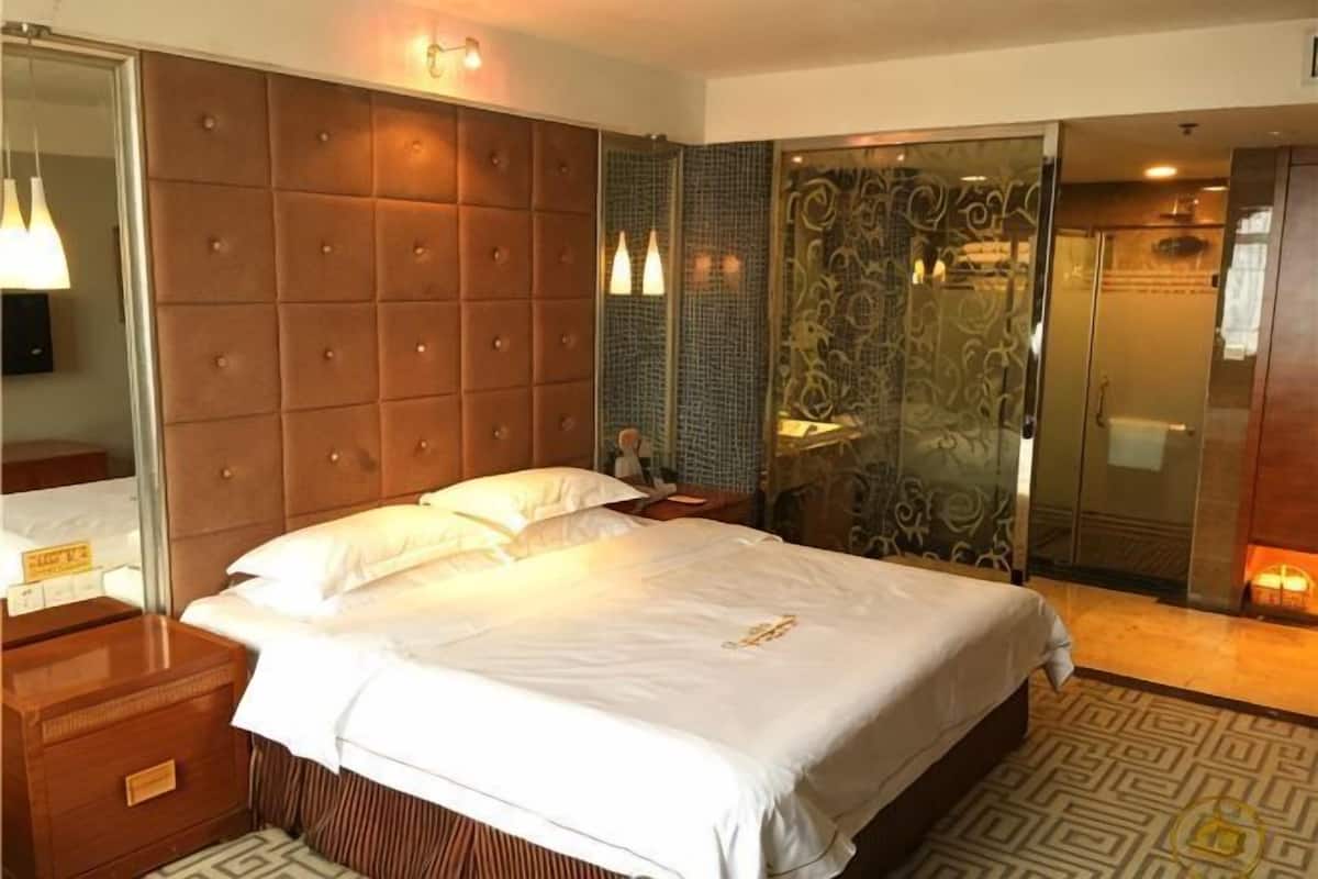 A hotel room with a large bed, a wooden nightstand, a mirror, a glass shower enclosure, and a patterned floor.