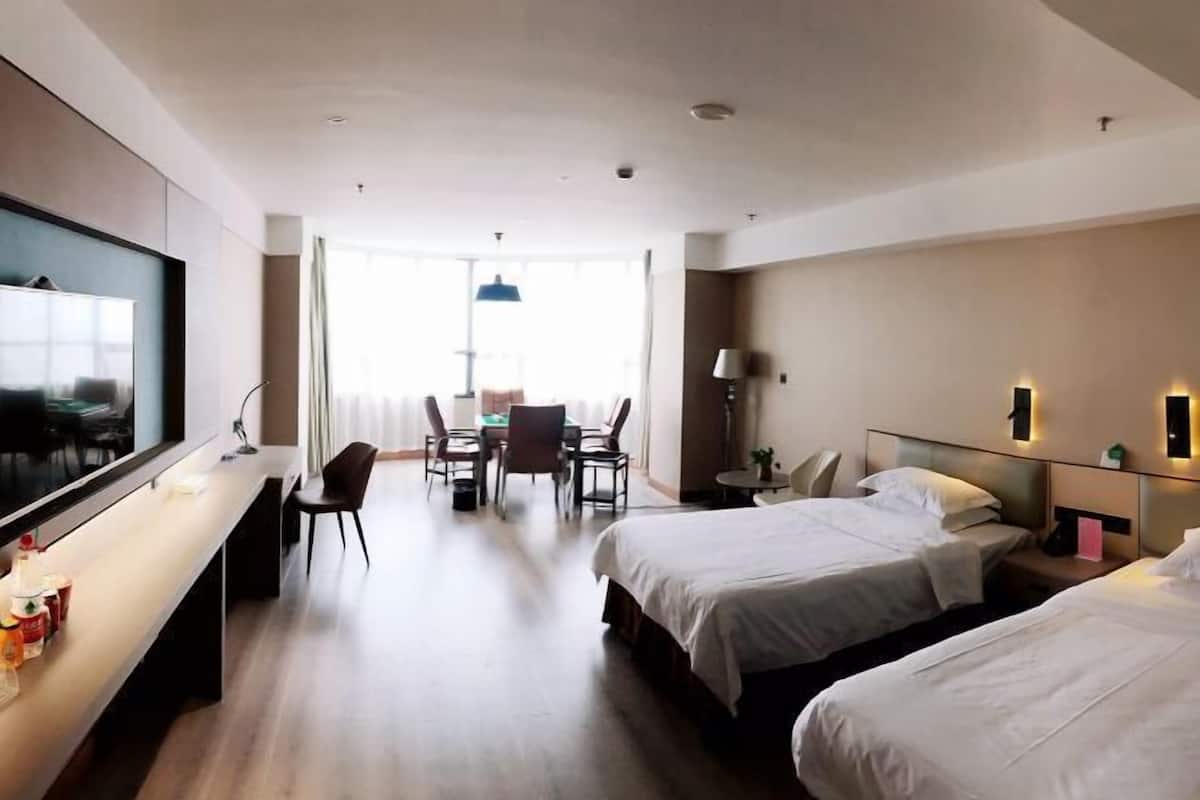 A hotel room with a TV, two beds, a desk with chairs, a dining table, and a lamp.