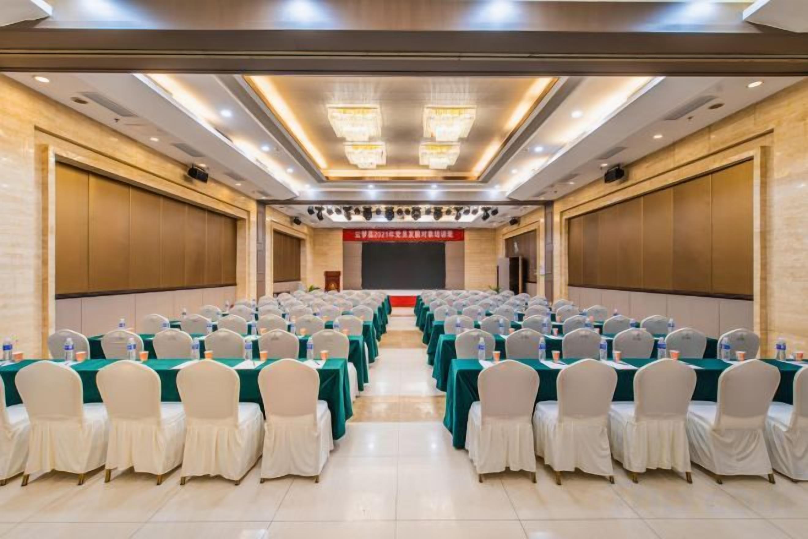 Un salón de conferencias con mesas y sillas dispuestas en filas, un escenario y un cartel rojo.