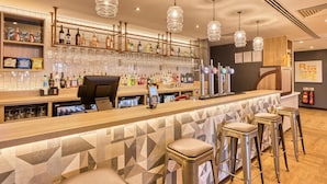 Un espace bar avec un comptoir, des tabourets, des luminaires suspendus et des étagères remplies de bouteilles et de verres.