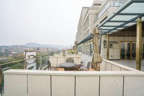 Una terraza en la azotea con una mesa y sillas, barandilla de vidrio y vistas panorámicas de la ciudad.