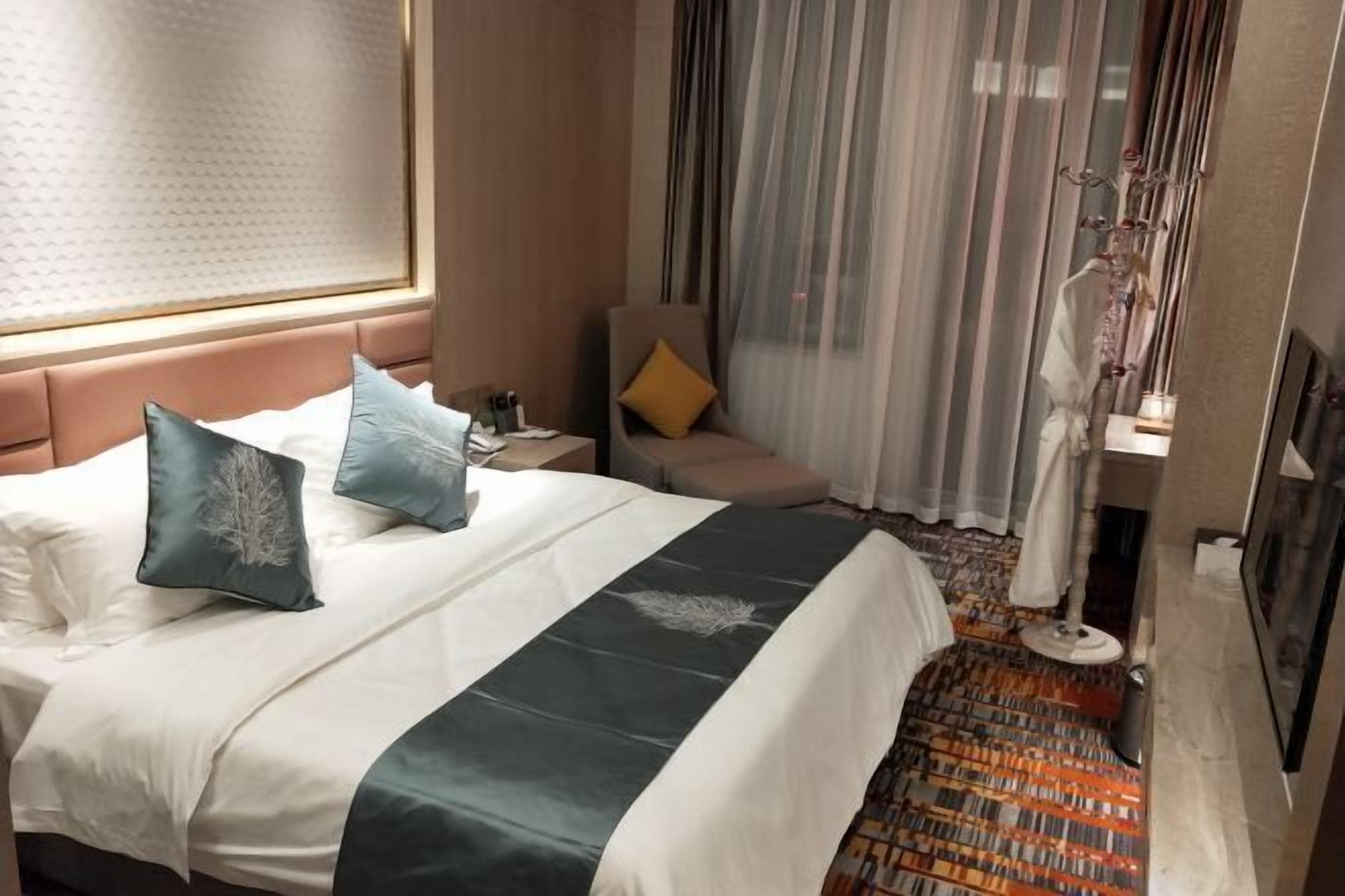 A hotel room with a bed, a nightstand, a chair, a window with curtains, and a floor lamp.