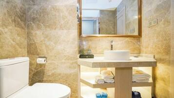 A bathroom with a marble wall, a toilet, a sink with a mirror above it, and shelves with towels and slippers.
