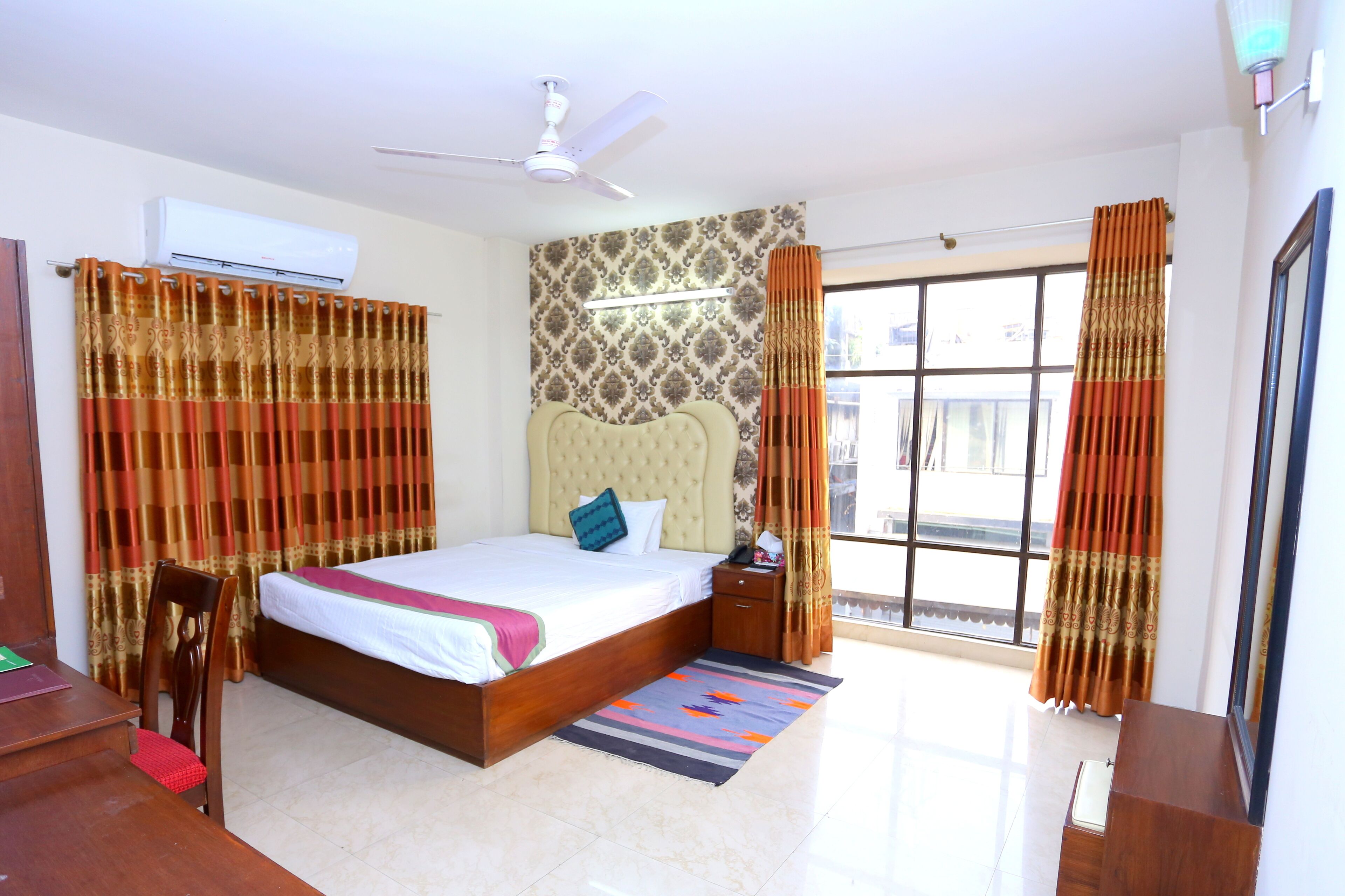 A bedroom with a bed, a nightstand, a chair, a desk, a window with curtains, and an air conditioning unit.